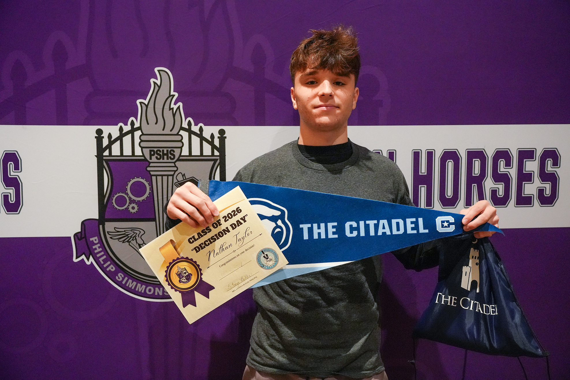 A person holds a certificate and a pennant in front of a purple backdrop with a logo.