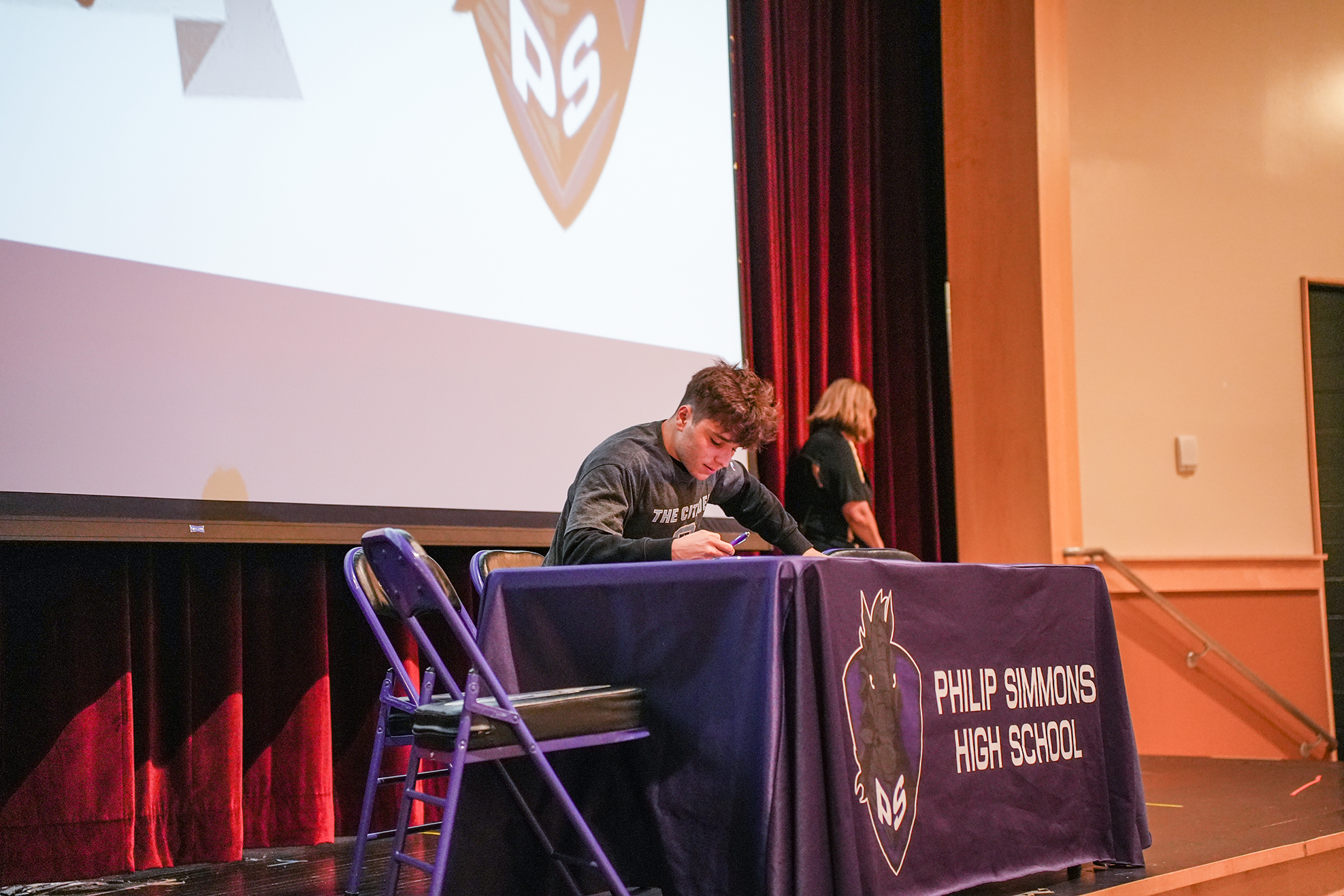 A person signing at a table with a purple cloth, logo, and text on it on a stage.