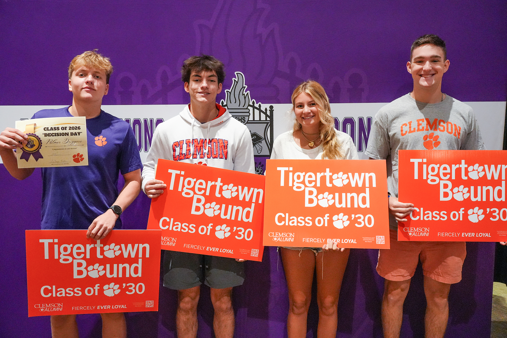 Four people stand in a row holding orange signs reading "Tigertown Bound Class of 2030." They are in front of a purple background.