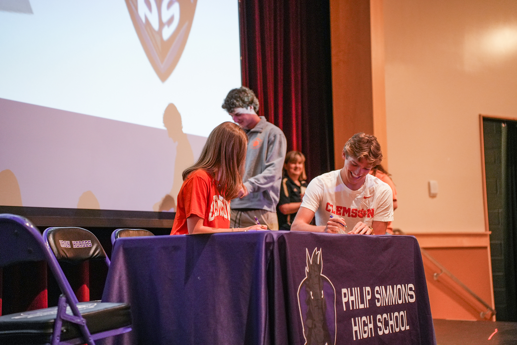 Three people sit at a table with a banner, one signing a document. Behind them, a projector screen displays an image.