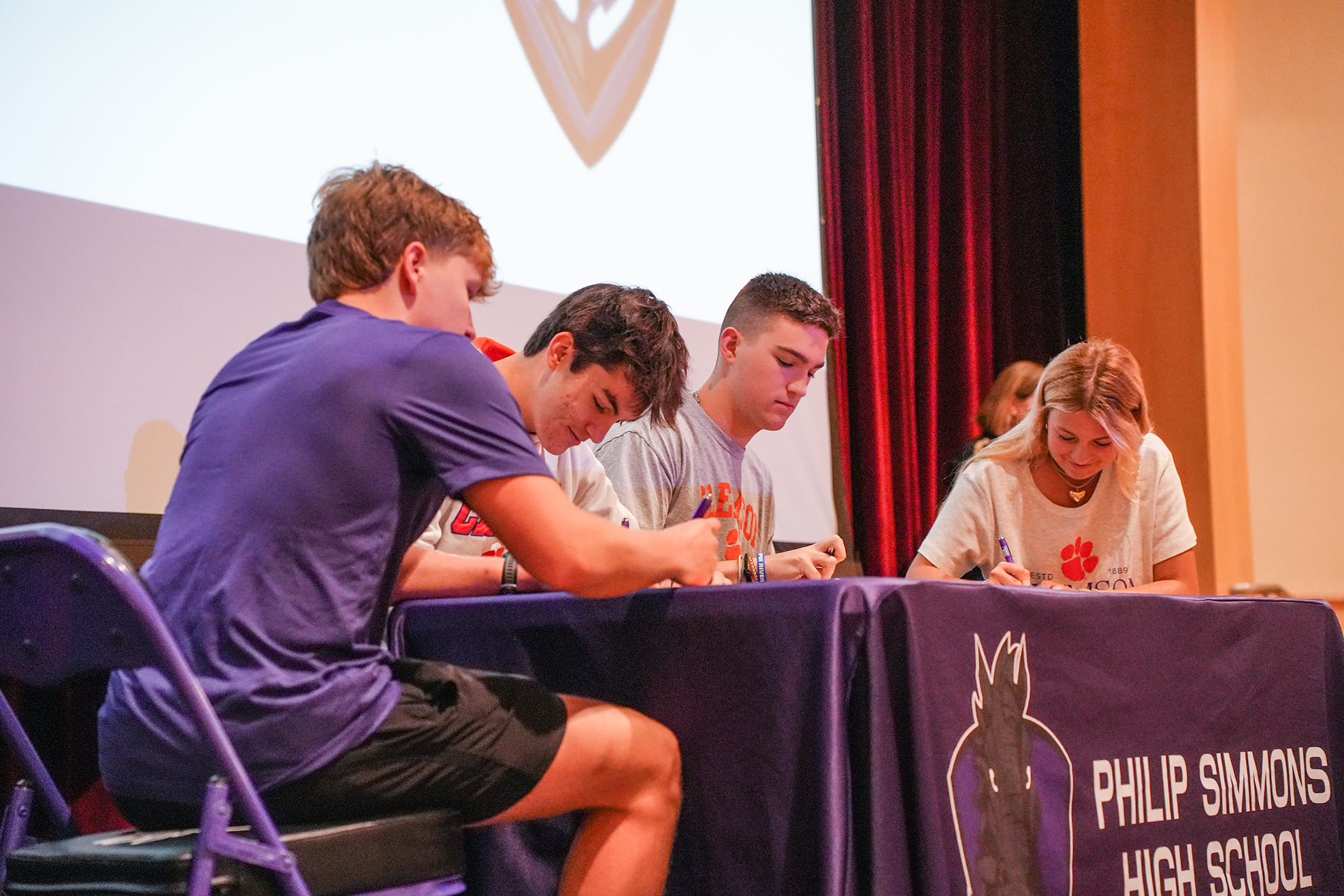Four people sit at a table signing papers. The table has a purple tablecloth with Philip Simmons High School logo.