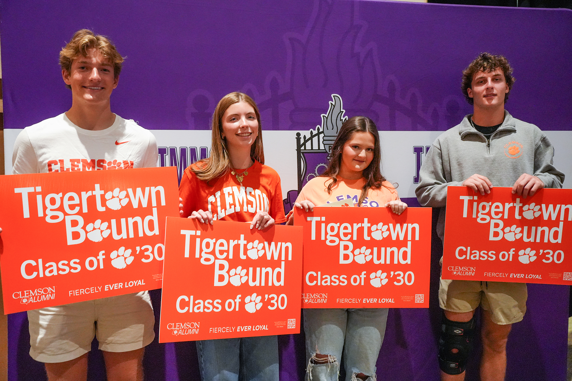 Four people hold red signs with "Tigertown Bound Class of '30" in front of a purple background.