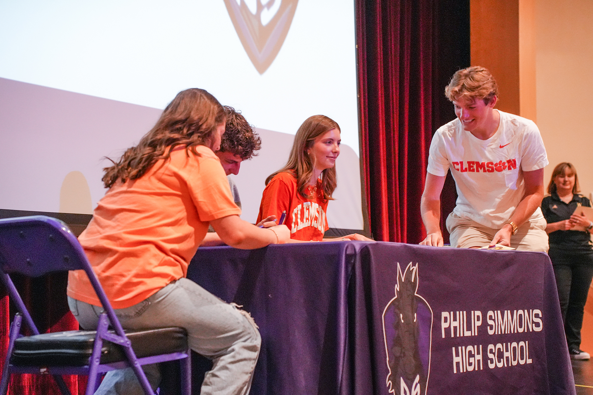 Students at a signing event wearing orange shirts. A table displays "Philip Simmons High School" and a logo.