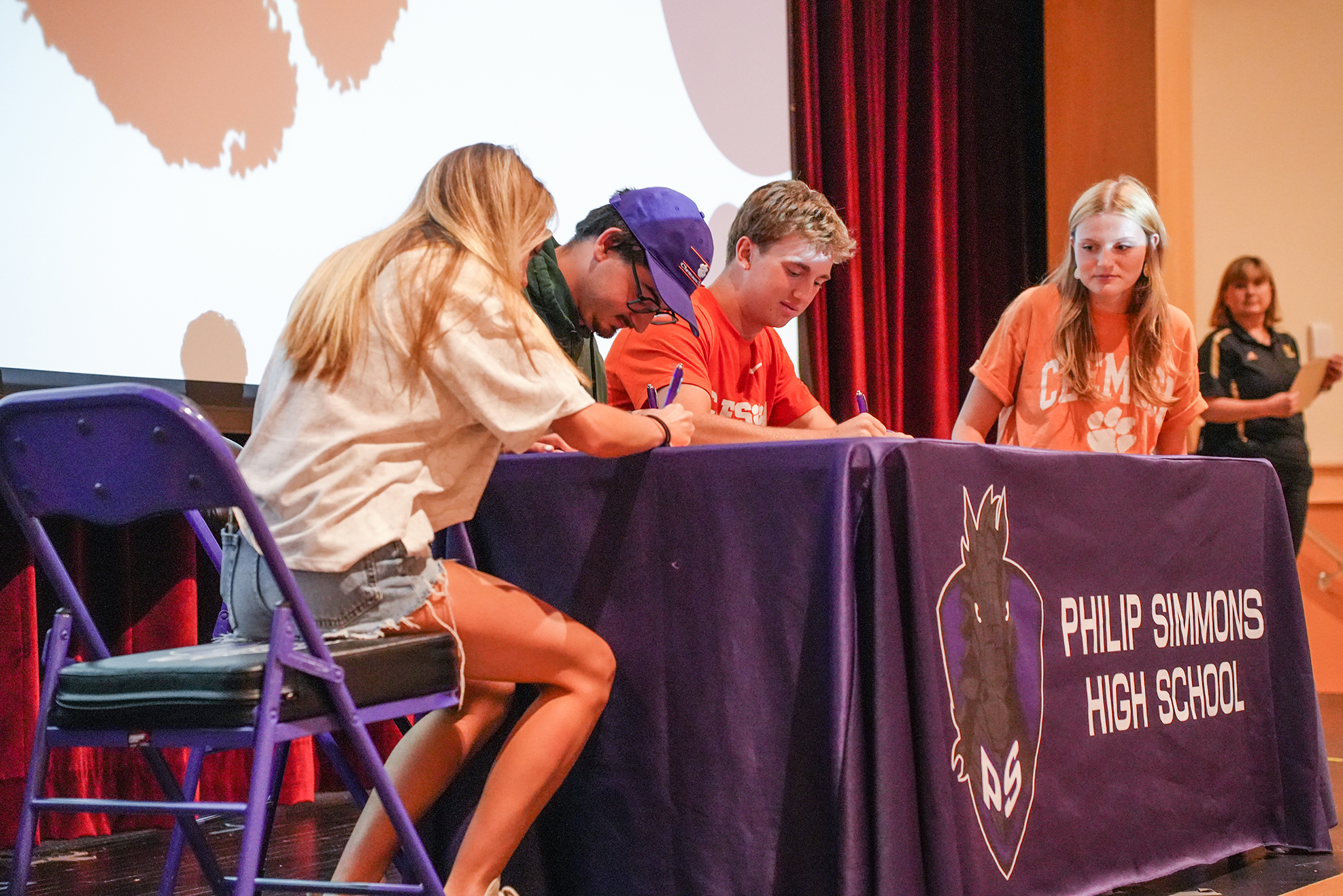 Four individuals, seated, are signing a document at a table covered with a purple cloth.