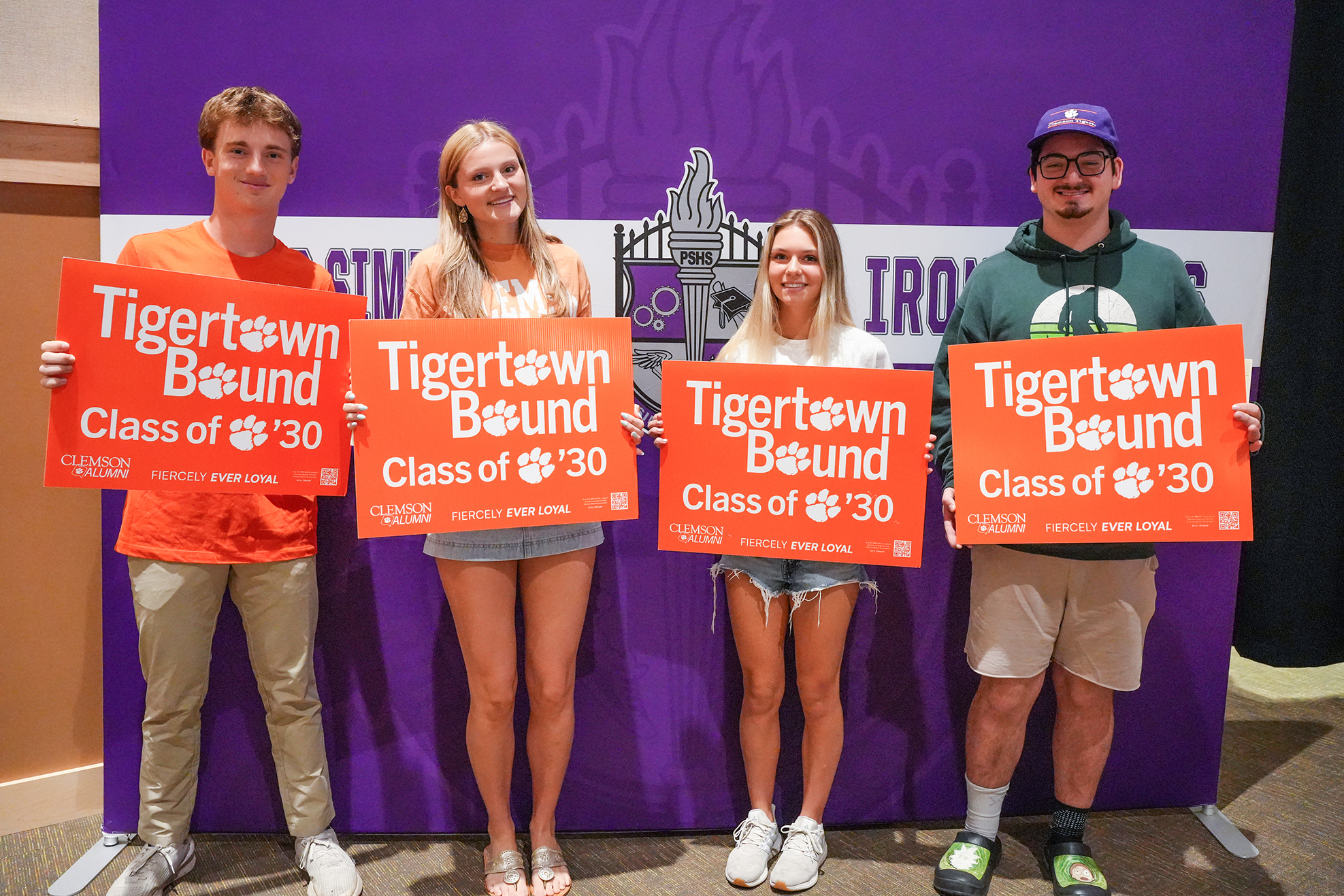 Four people stand before a purple backdrop, holding signs reading "Tigertown Bound Class of '30."