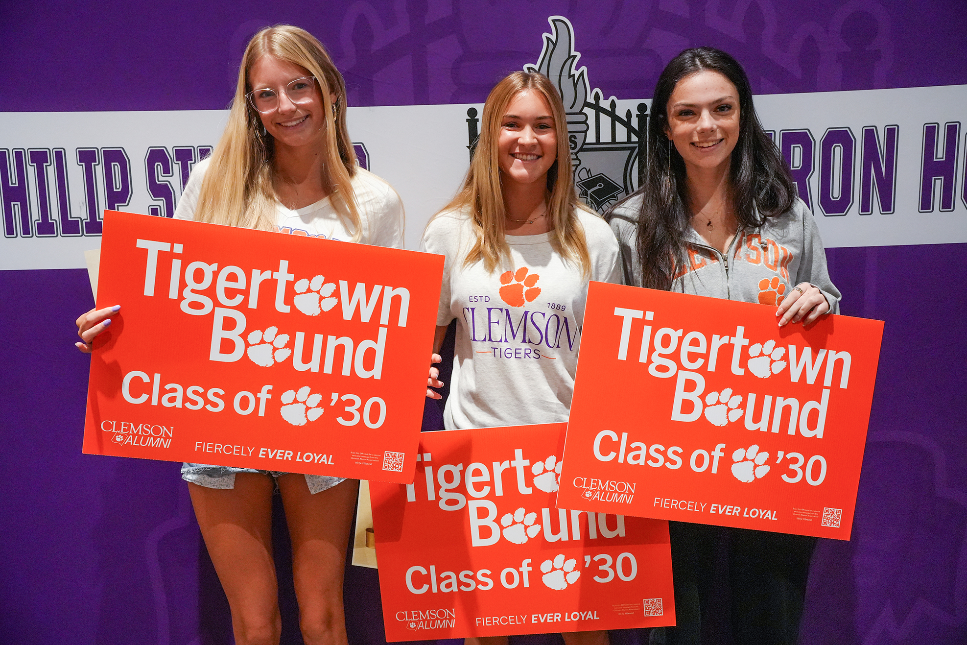 Three women in front of a purple background, holding red signs that say "Tigertown Bound Class of '30".