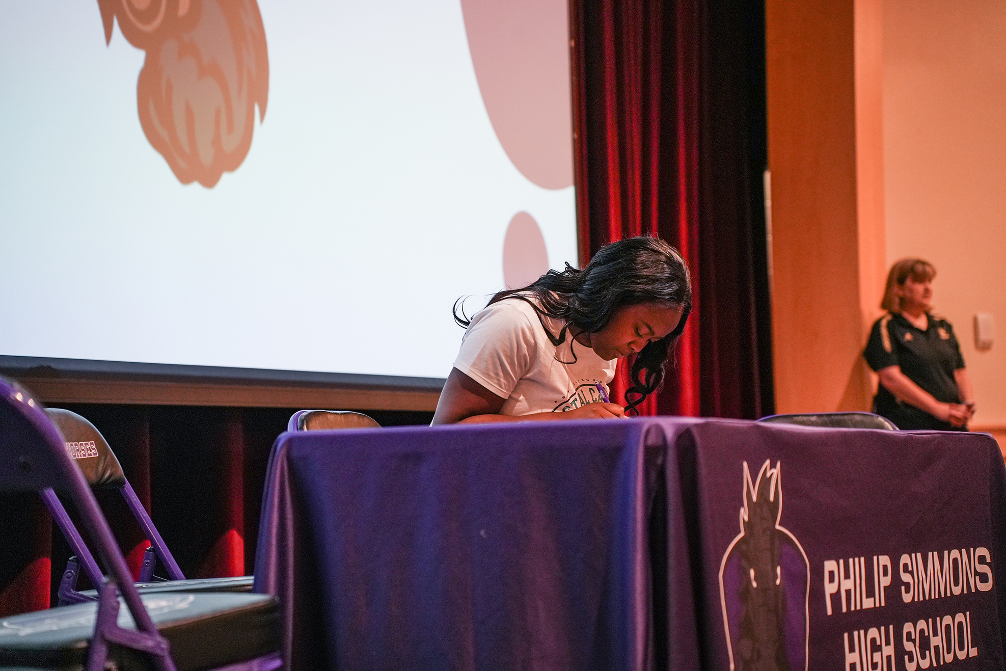 A woman in a white t-shirt sits at a purple table, writing, with a screen behind her.