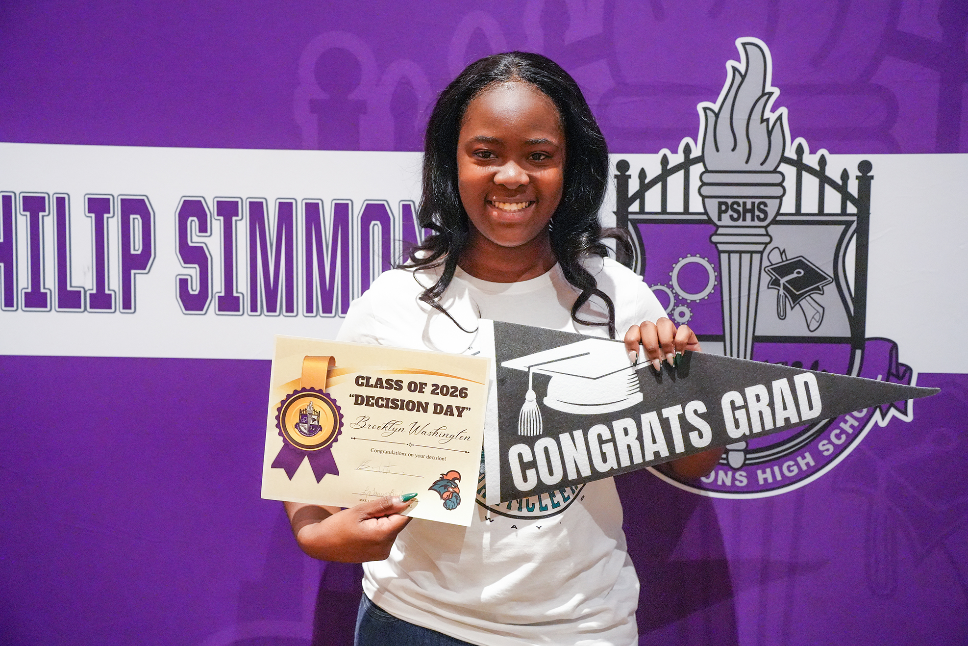 A person holds a graduation cap and a certificate with a school logo in the background.