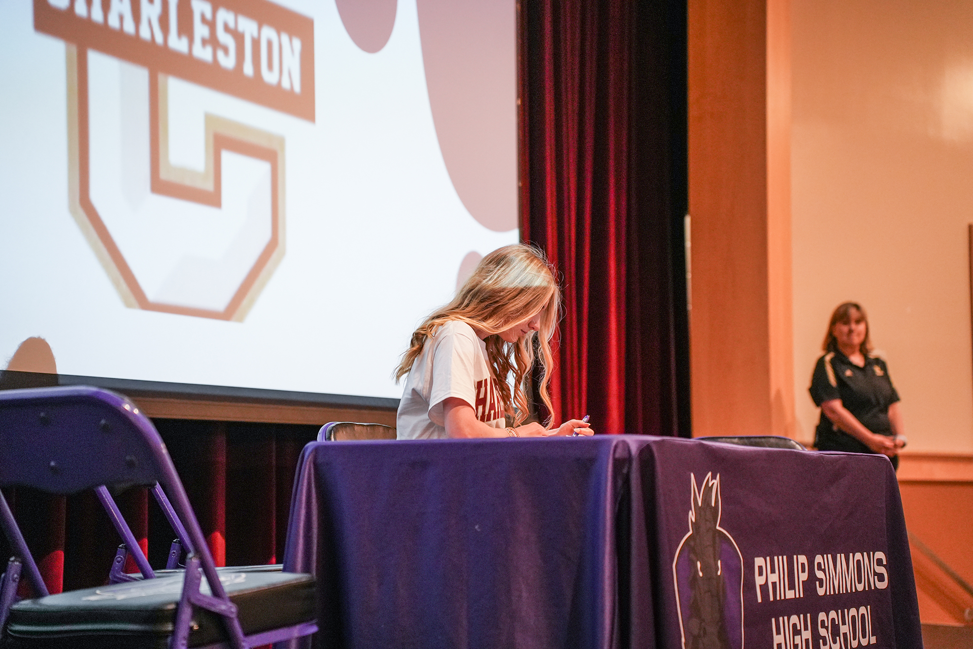 A student signs a document at a desk under a screen with the words "Charleston" and "Philip Simmons High School".