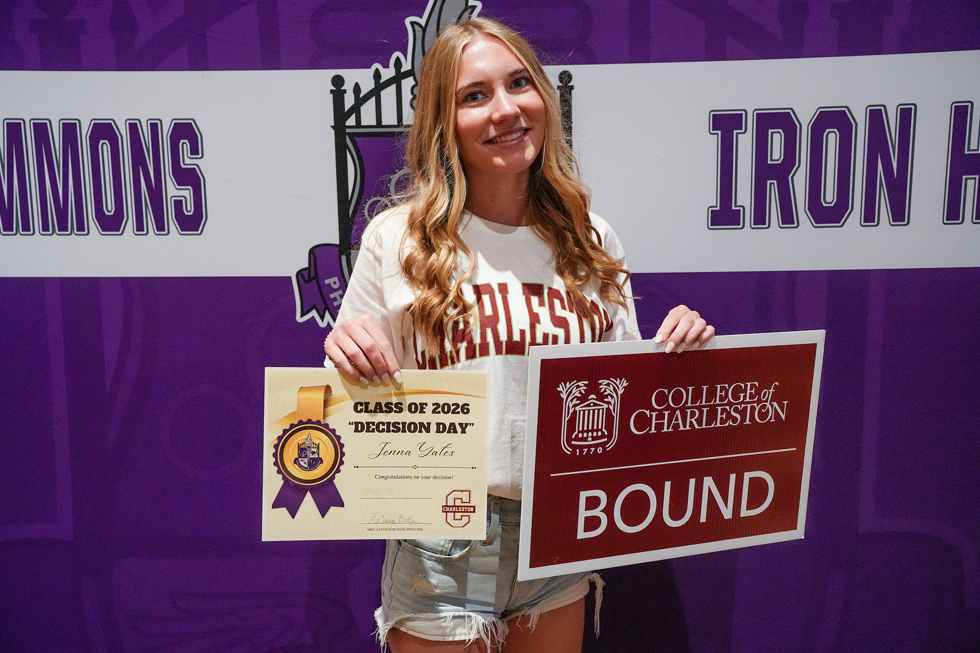 A young woman in a white shirt stands against a purple backdrop, holding a sign reading "Bound" and a certificate.