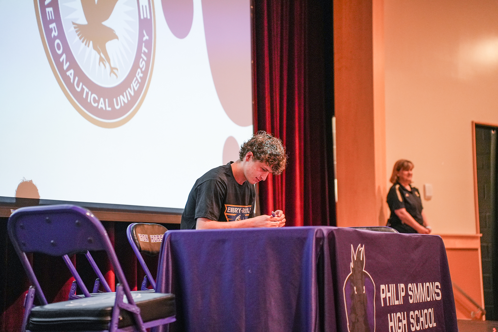 A person is seated at a table, signing papers. Behind them is a screen with a logo.