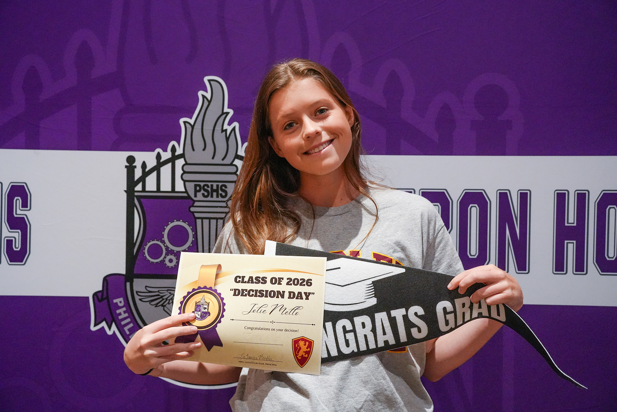 A smiling girl holds a certificate for "Class of 2020 Decision Day" and a graduation sash against a purple backdrop.