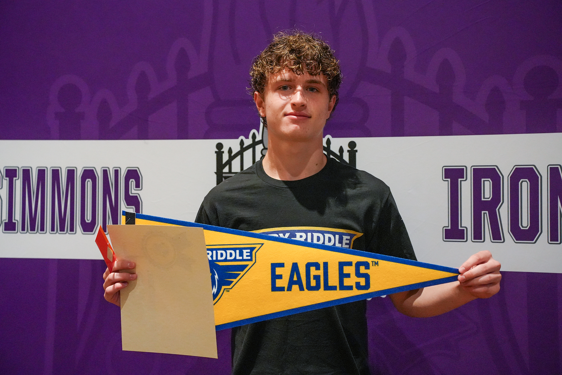 A man stands in front of a Simmons Iron banner, holding a pennant reading Eagles. He wears a black T-shirt.