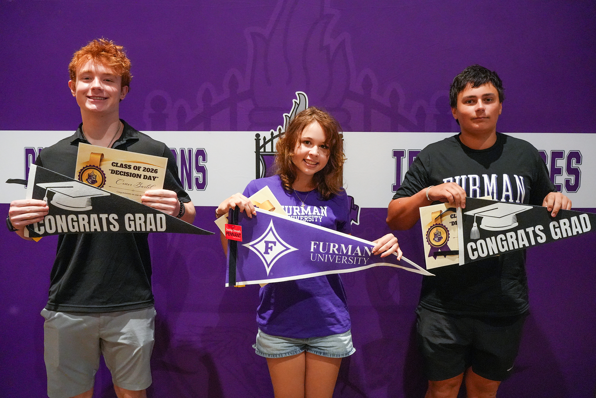 Three people pose with graduation caps and diplomas against a purple backdrop with university logos and banners.