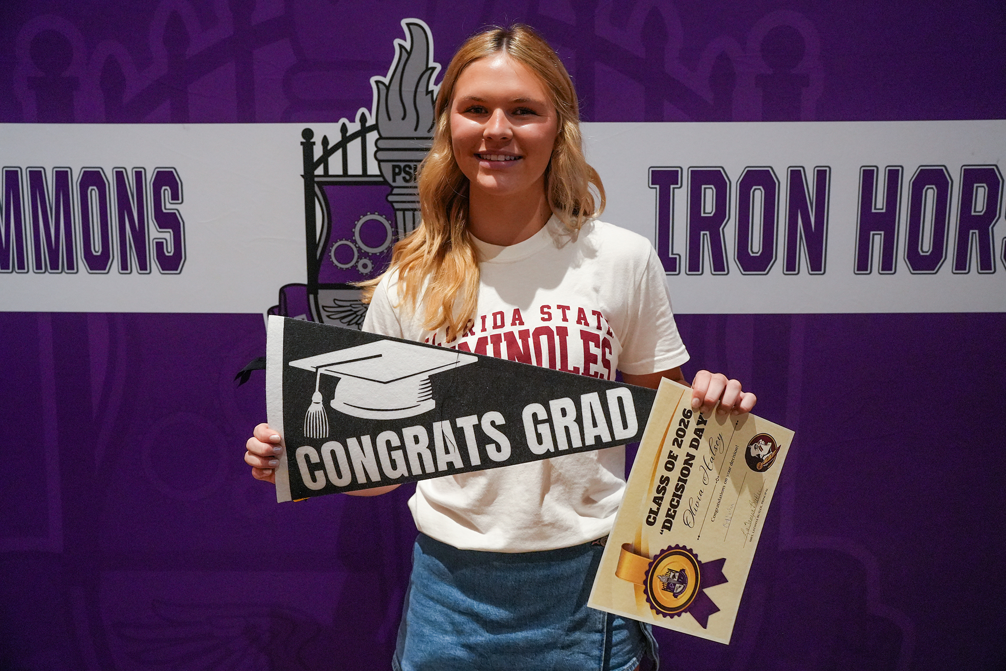 Woman in white t-shirt holds graduation cap sign and certificate in front of purple backdrop.