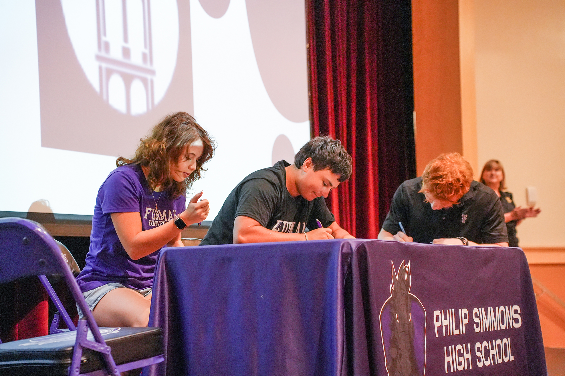 Four students sit at a table signing papers. A large projector screen is behind them.