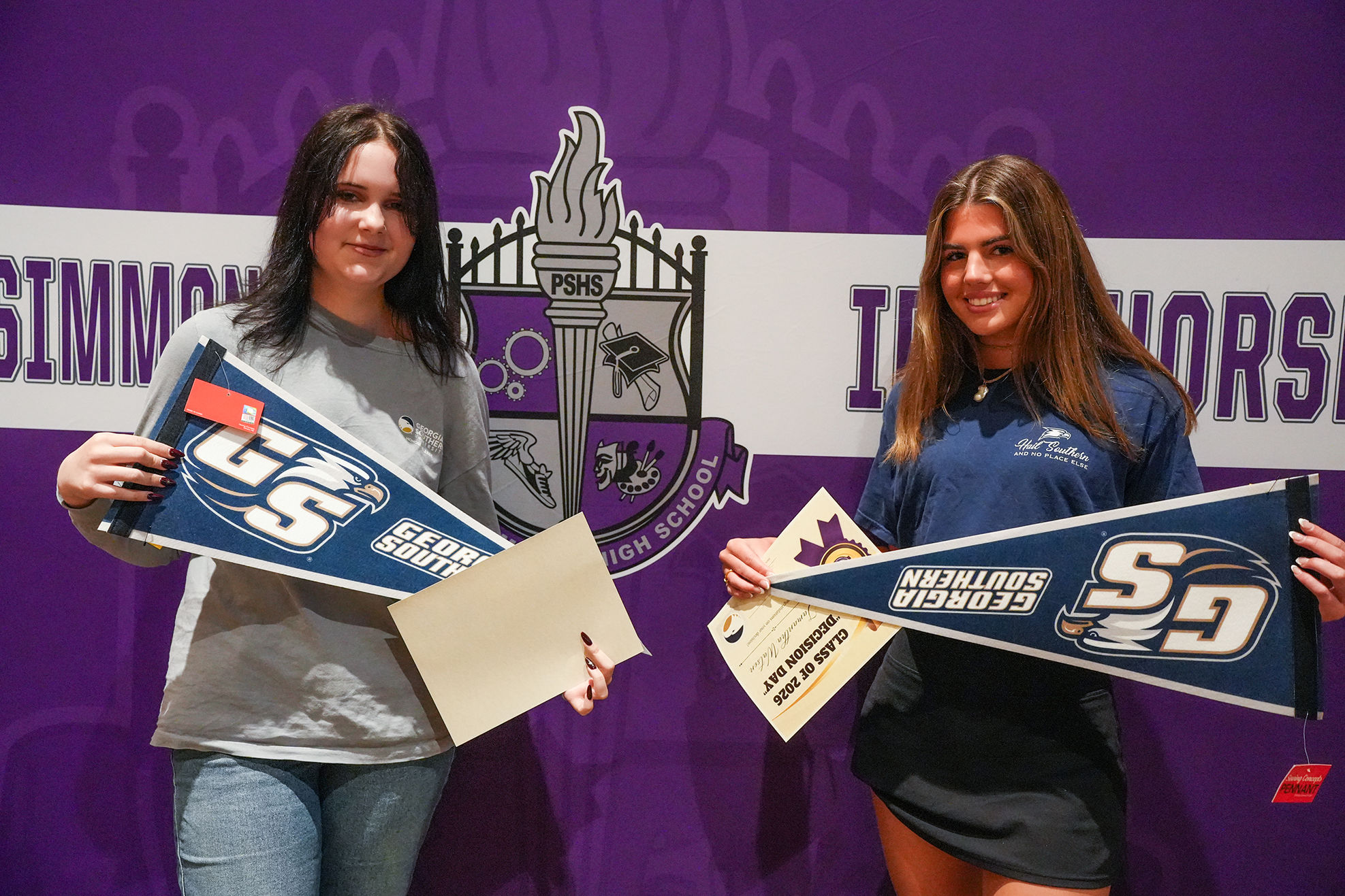 Two women hold pennants labeled "Seniors" in front of a purple backdrop with a school emblem.