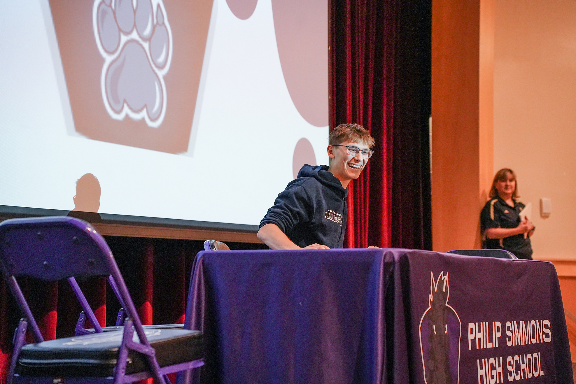 A person sits at a table in front of a screen displaying a paw print. Another person stands at the back.