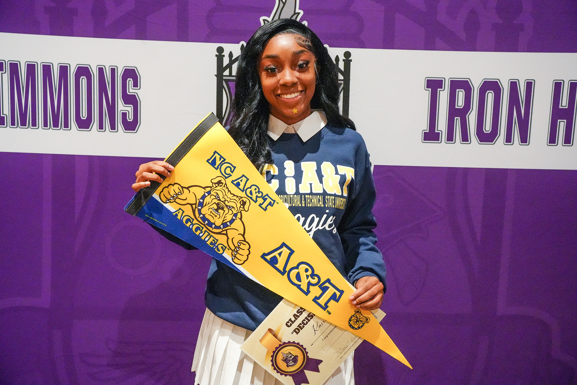 Woman holding a pennant and diploma stands in front of a purple backdrop with a white sign.