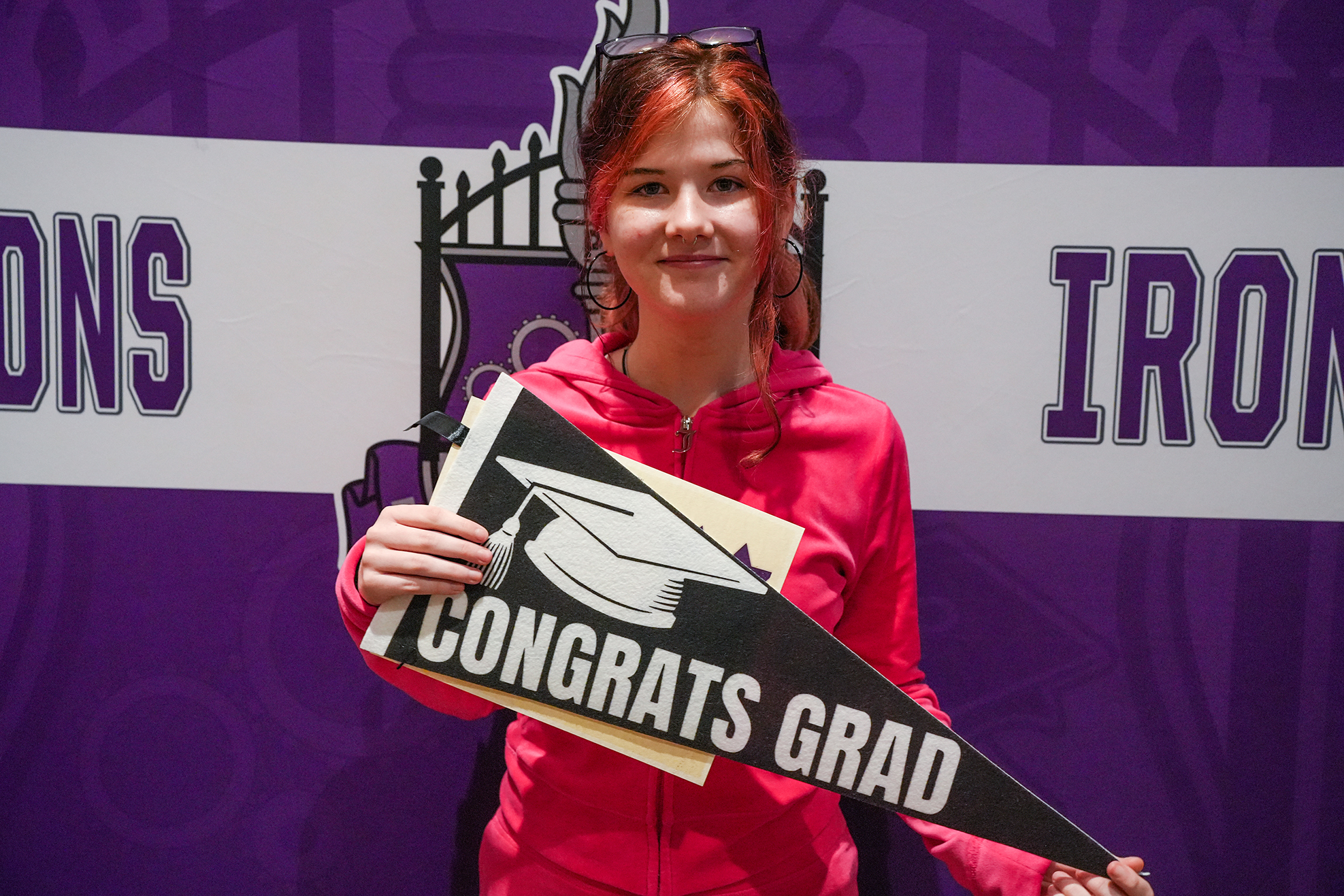 A smiling woman in red holds a graduation cap banner in front of a backdrop reading "Irons" and "Irons."