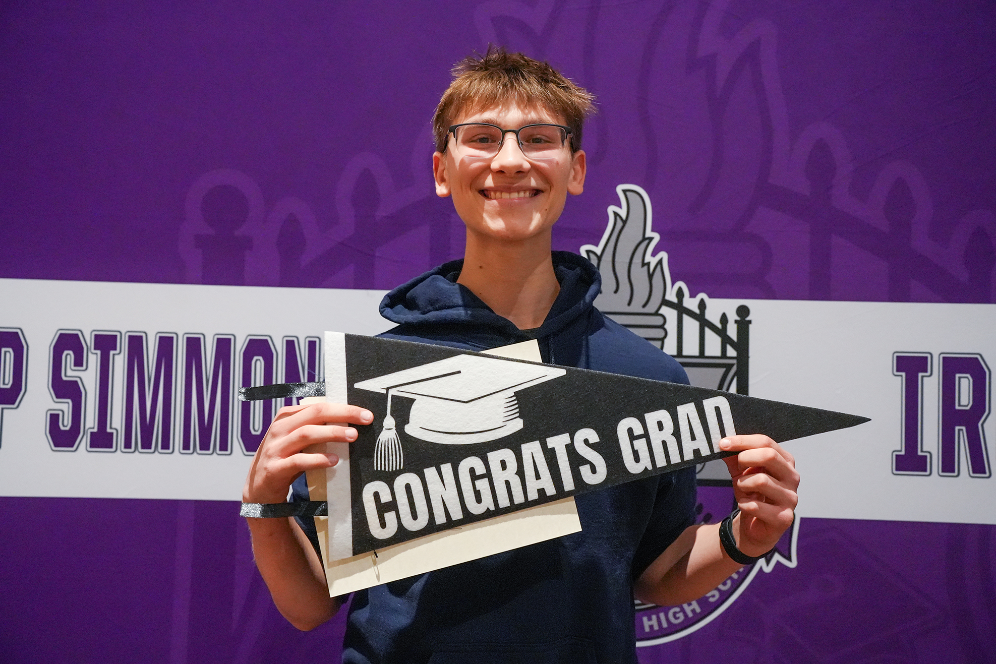 A person in a navy blue hoodie holds a graduation cap banner with the words "CONGRATS GRAD" in front of a purple backdrop.