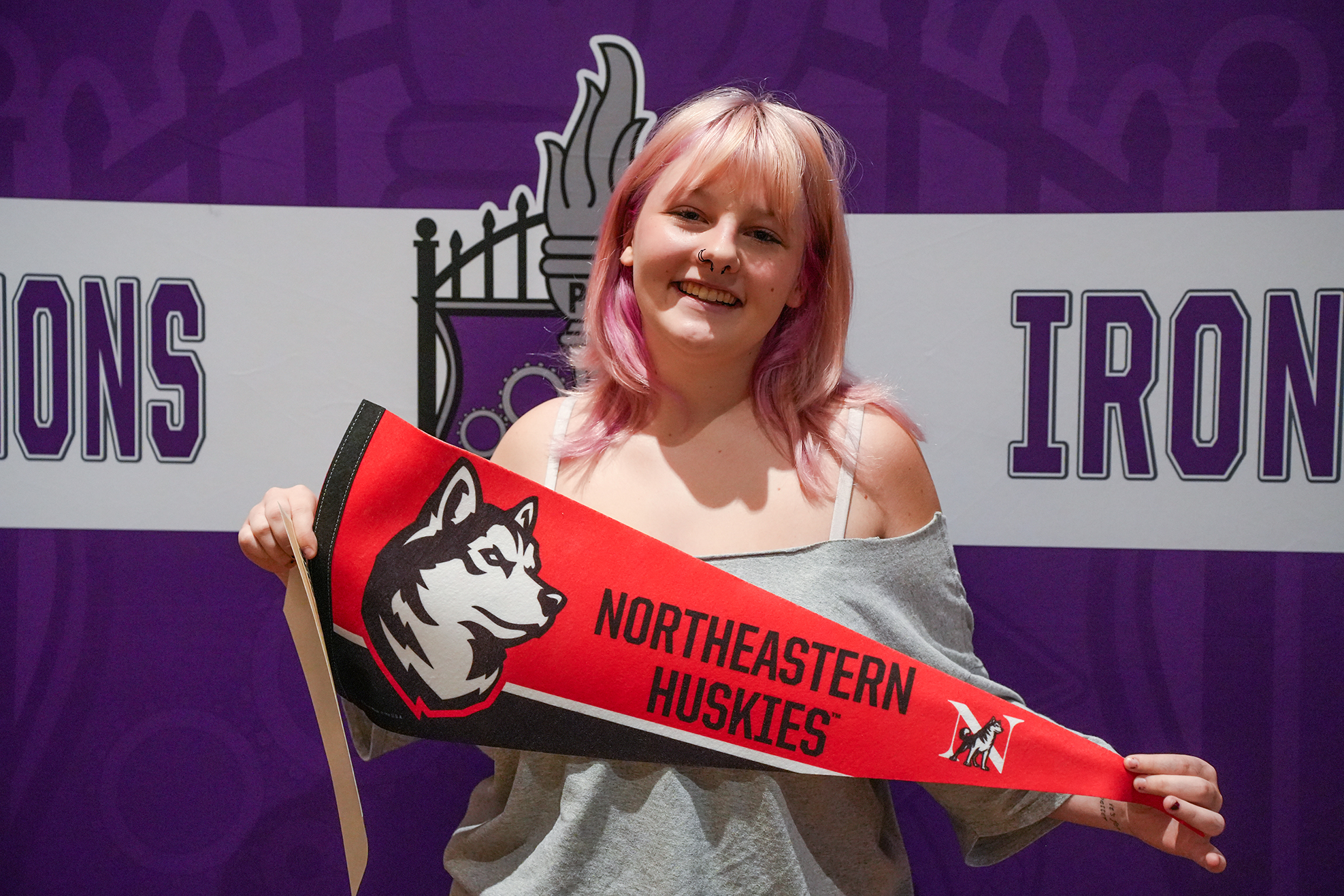 A woman with pink hair holds a Northeastern Huskies pennant banner in front of a purple backdrop.