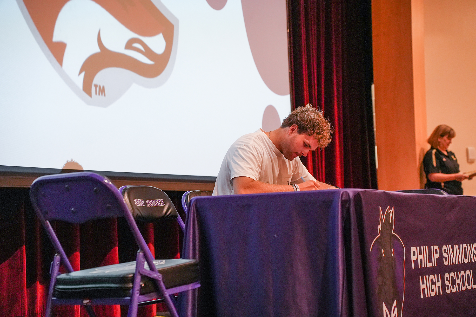 A person sits at a table, writing on paper in front of a screen with a logo.
