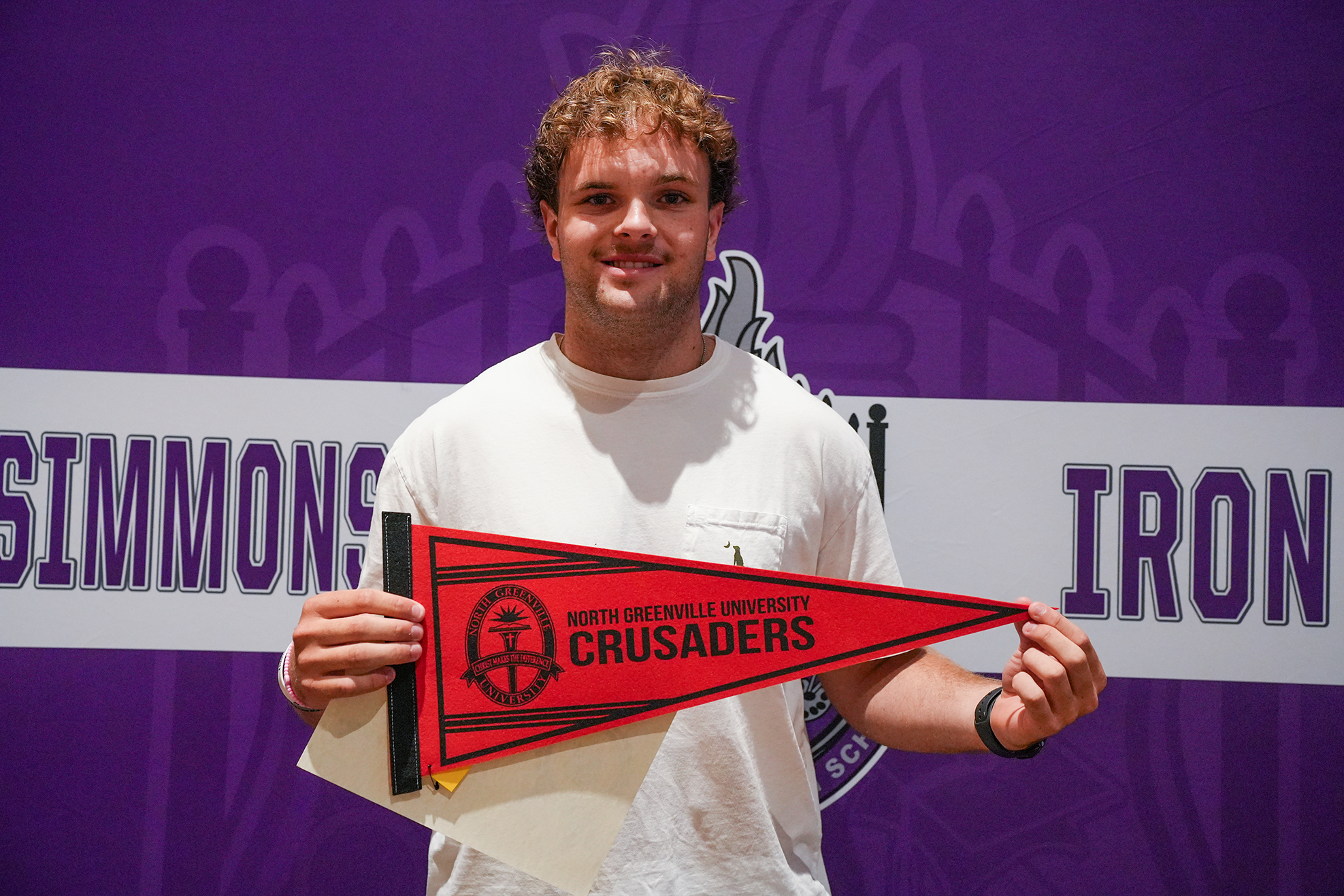 A man wearing a white T-shirt holds a red pennon banner reading "North University Crusaders" against a purple backdrop.