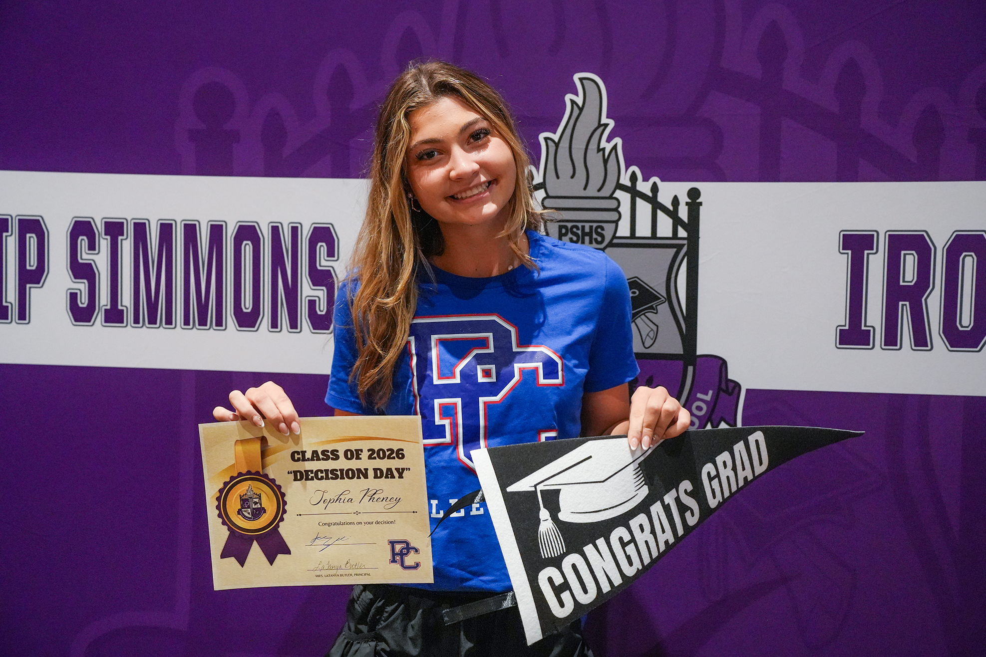 A woman poses with a certificate and a graduation banner. Behind her is a purple backdrop with "IP SIMMONS" and "IRO".
