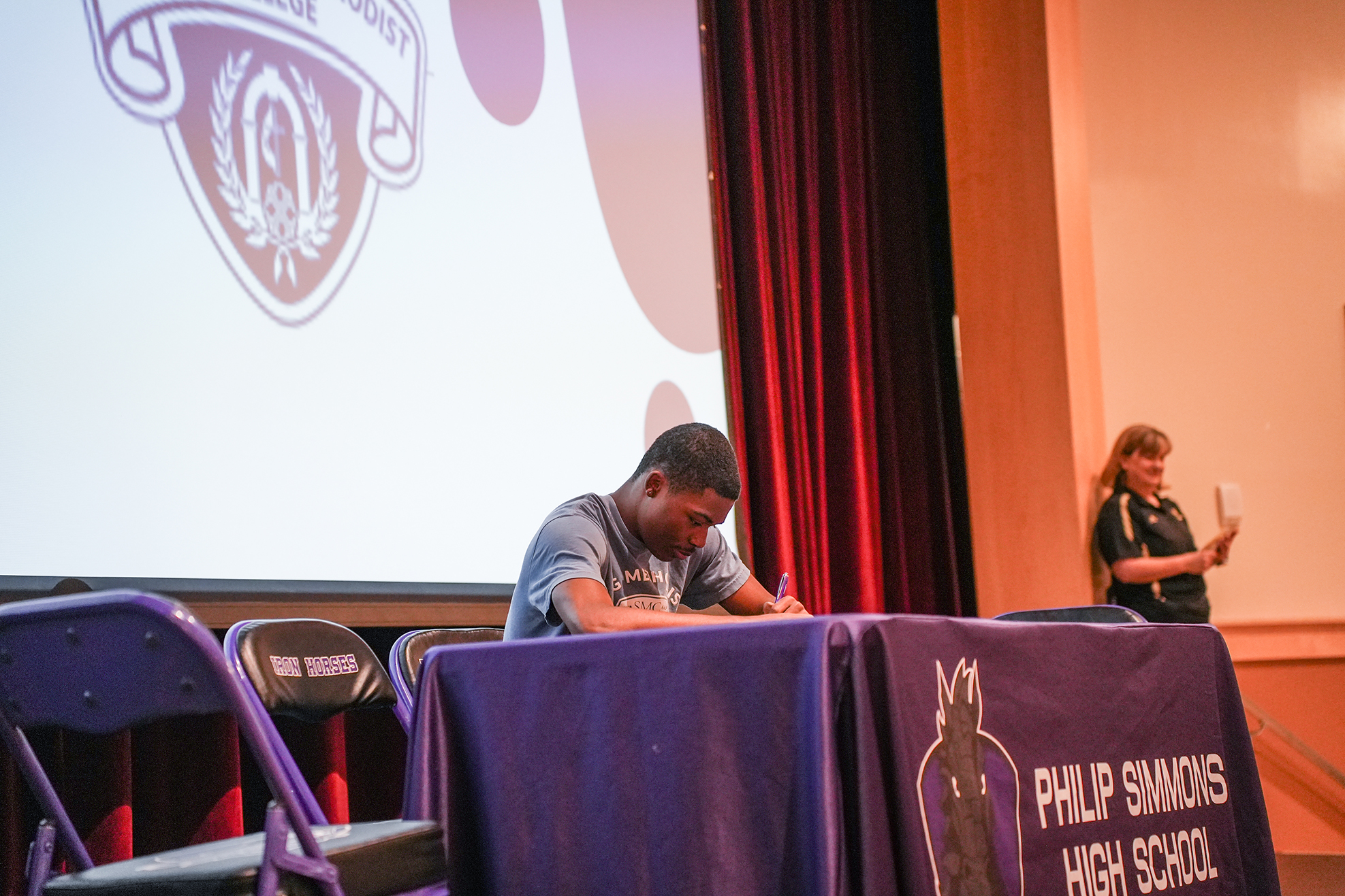Person at a table signing papers, a large screen behind them. A woman stands at the side.