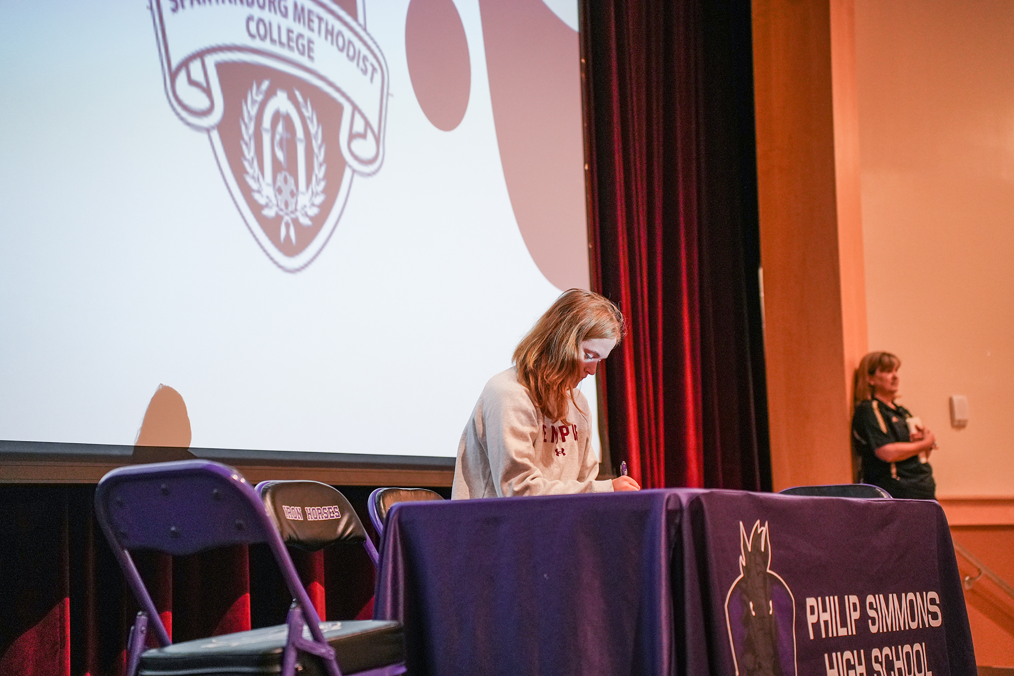 A woman sits at a table, writing, while another stands behind her. A projector screen shows a logo.