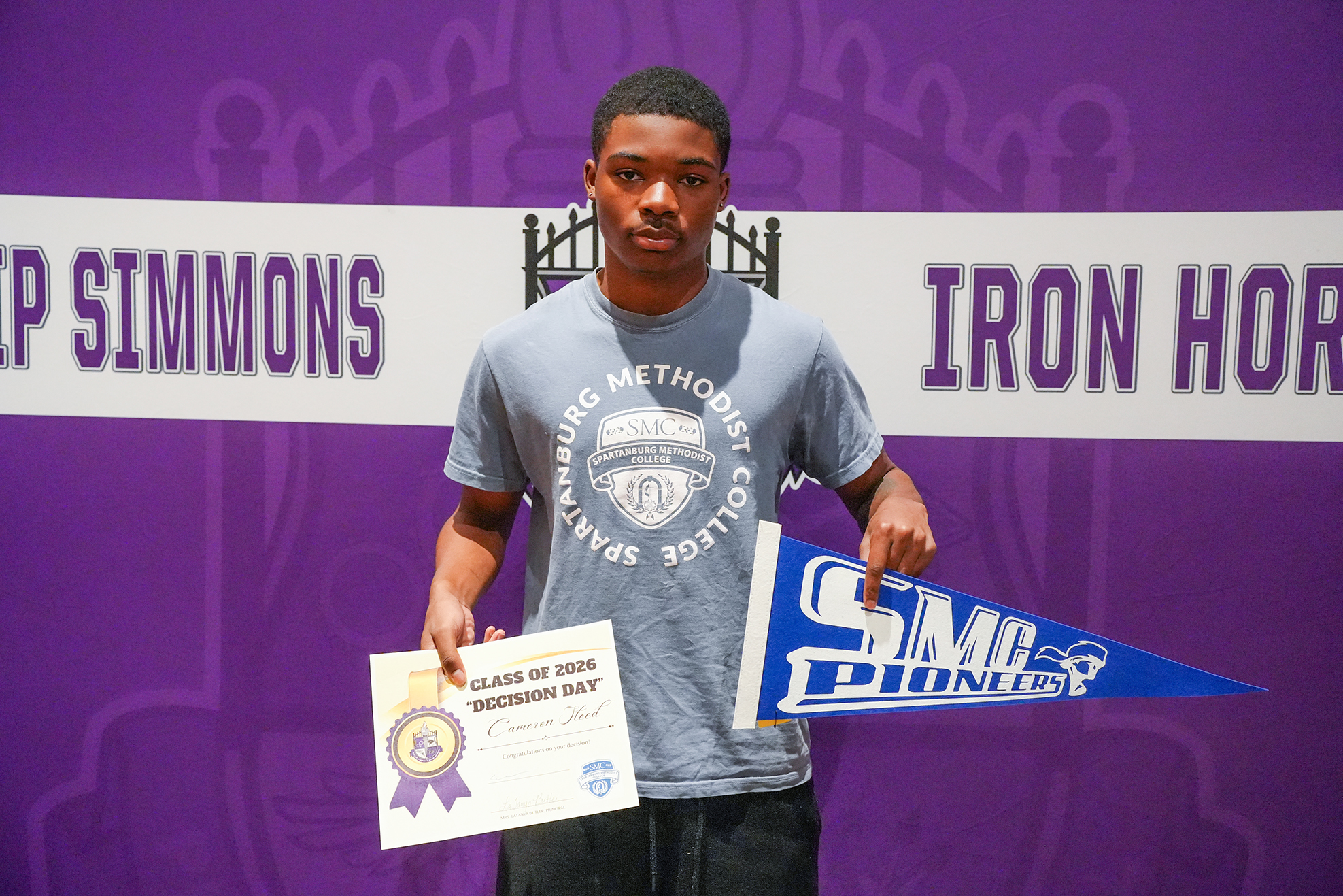 Person in gray T-shirt holding certificate and blue pennant with text. Purple backdrop displays "Iron Horse" and "Simmons" with logo.