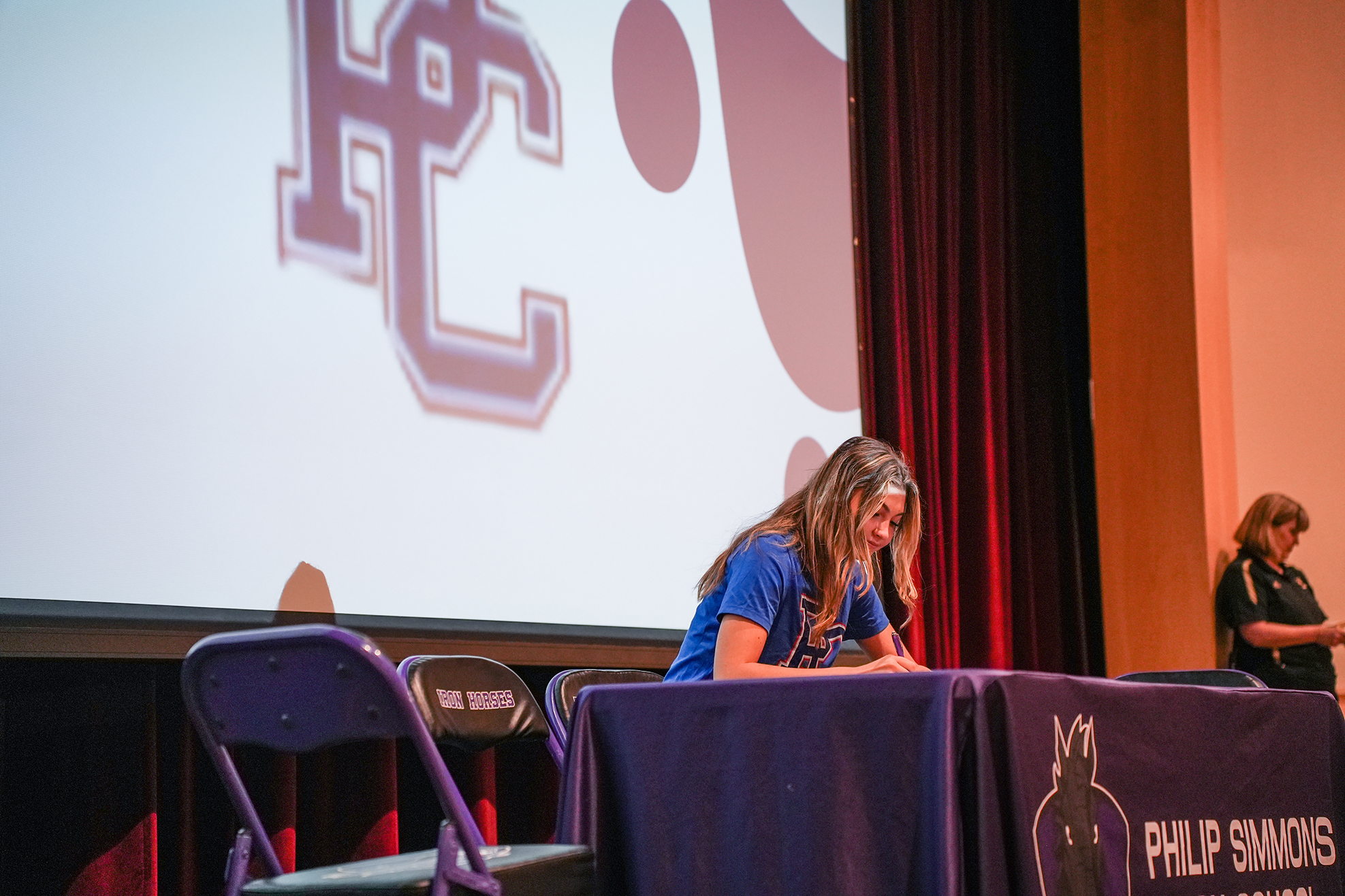 Woman in blue shirt signing an autograph on a table with a large logo behind her.