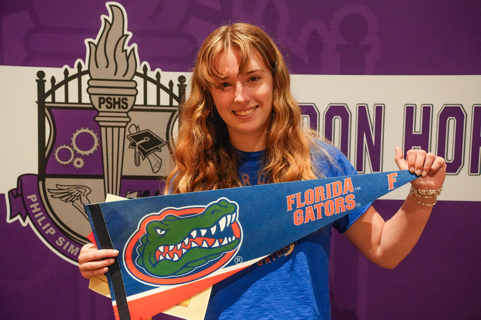 Woman with wavy hair wearing a blue shirt holds a Florida Gators pennant in front of a purple background.