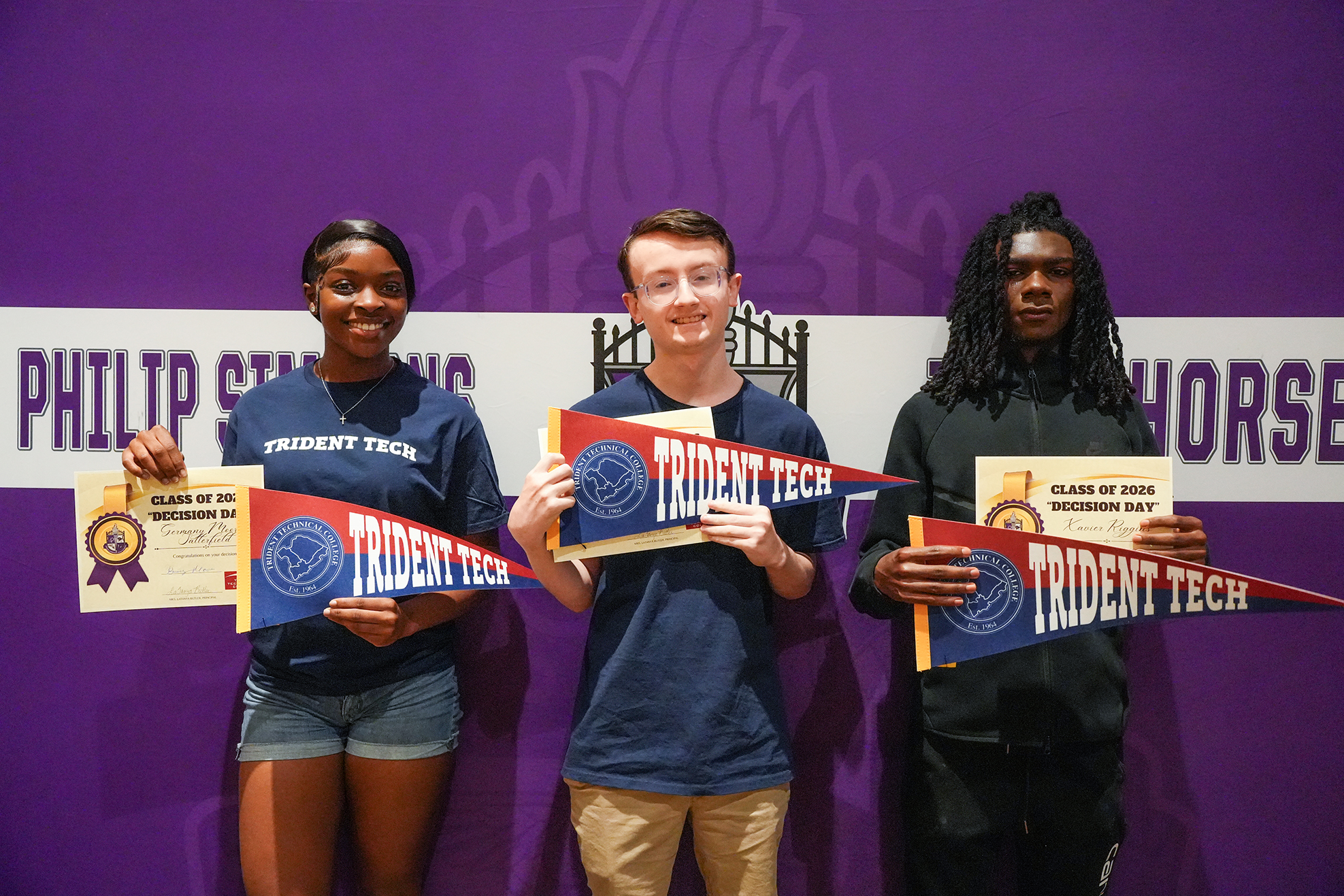 Three individuals pose, holding blue and red banners and certificates. One has a watch, and the wall behind them is purple with text.