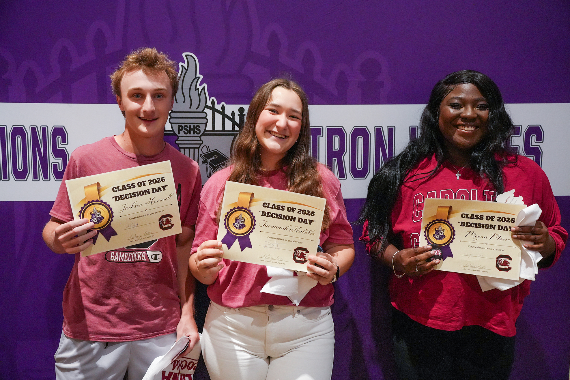 Three students, holding certificates, smile in front of a purple wall with "TRONS" and "PK" on it.
