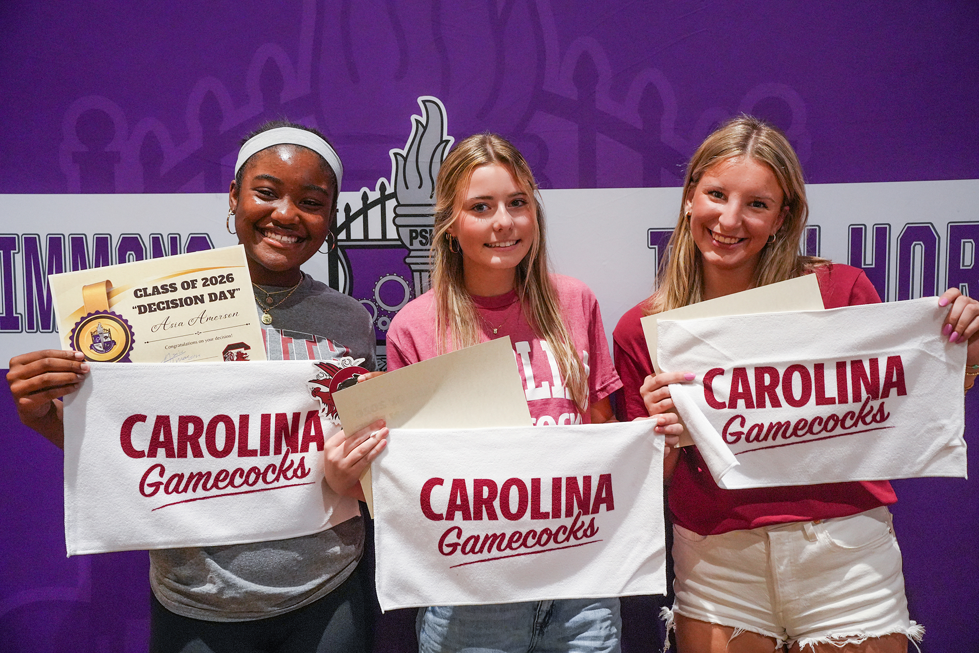 Three women hold diplomas and banners with "Carolina Gamecocks" text, standing in front of a purple backdrop with white text.