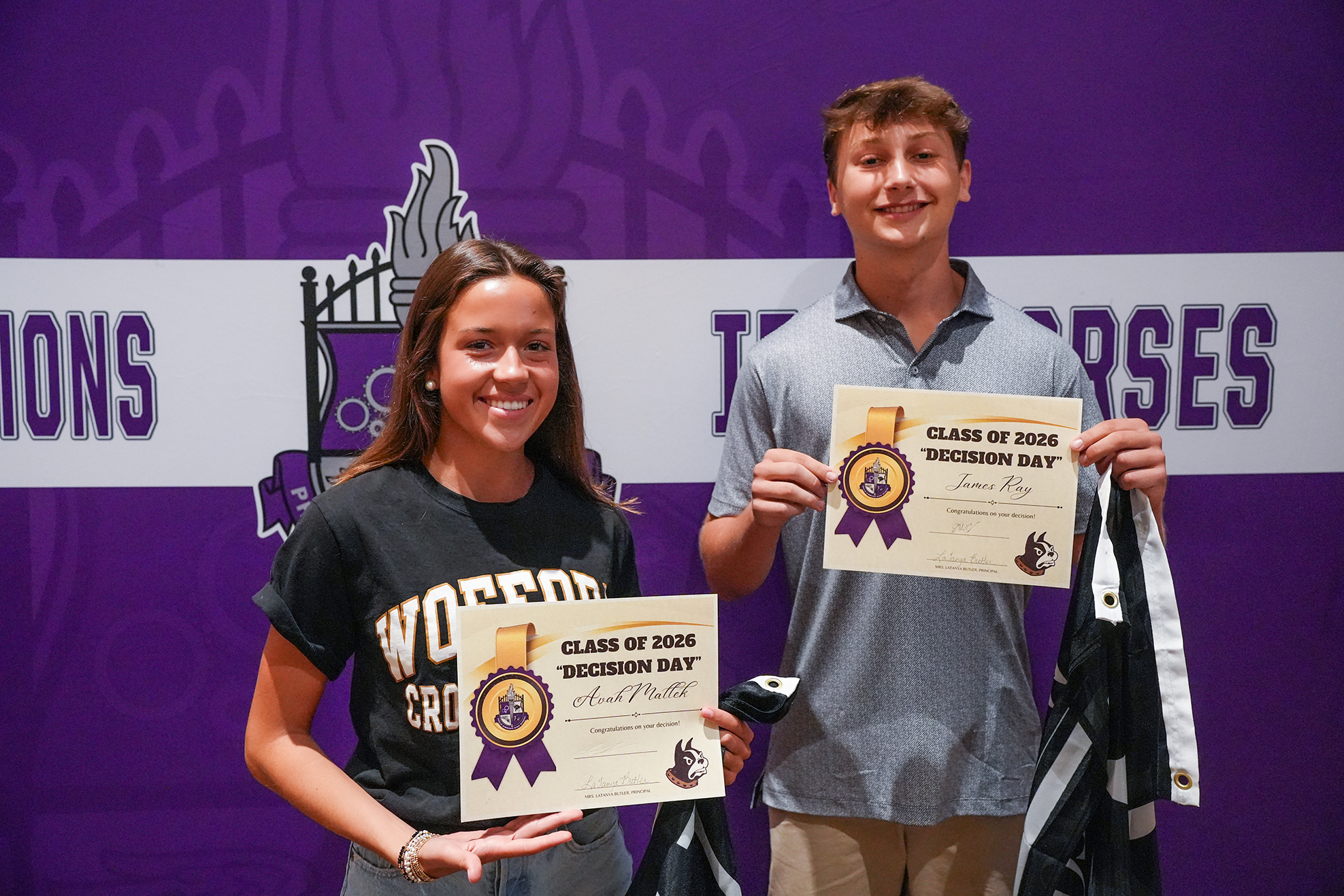 Two individuals stand before a purple backdrop with text and school emblems, holding certificates. The woman smiles, wearing a black shirt. The man grins, holding a certificate and jacket.
