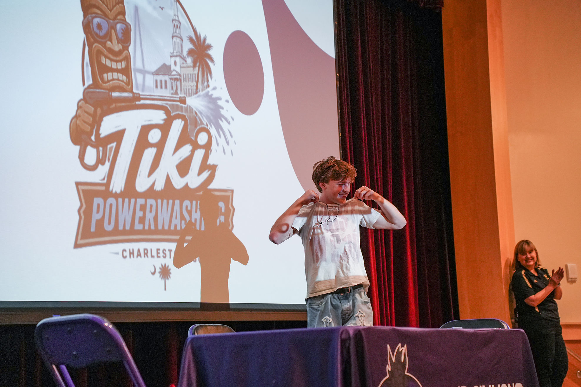 A boy stands at a desk in front of a screen displaying a logo and text. Behind him, a woman stands holding a camera.