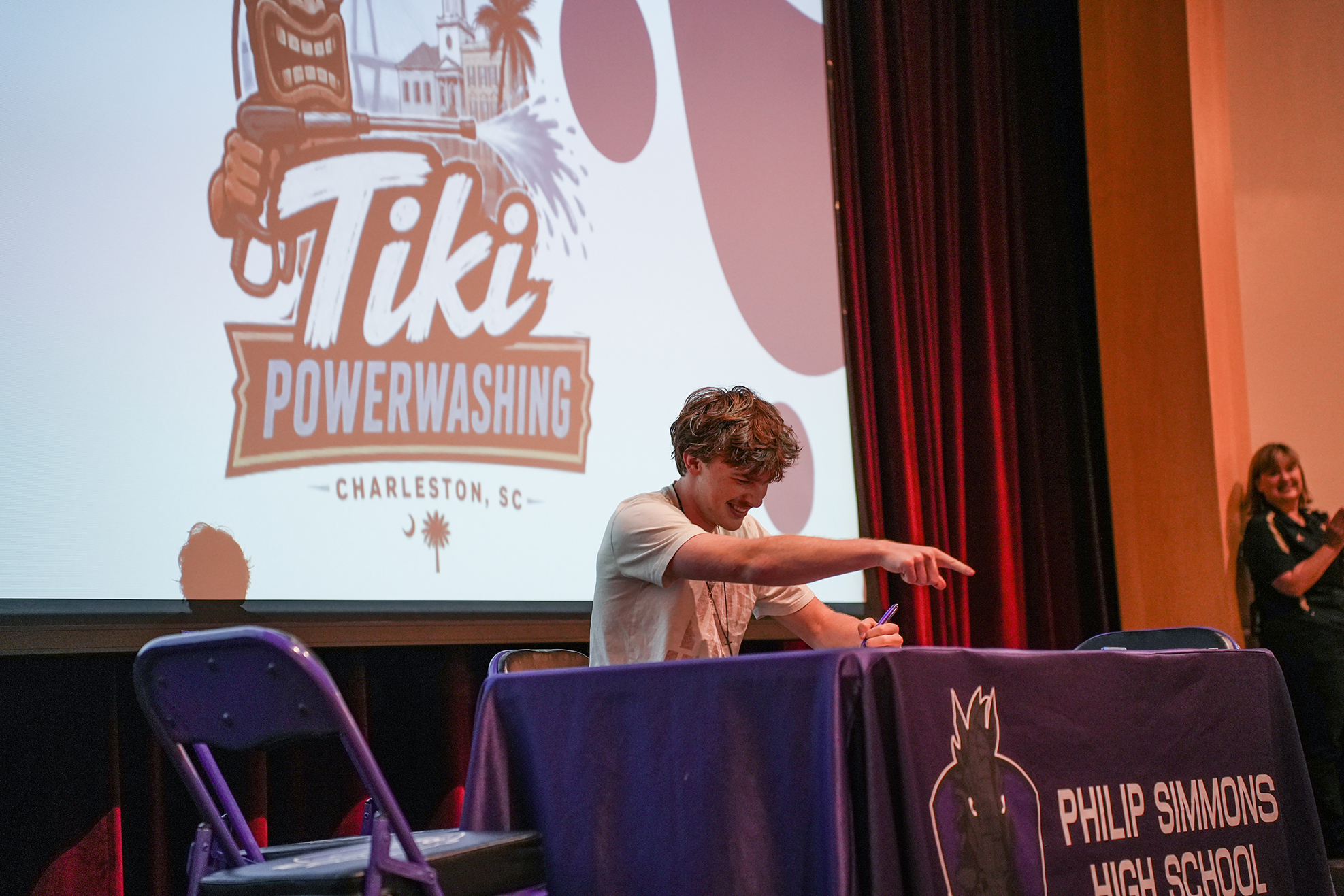 A man signs a table in front of a large Tiki Powerwashing banner, with a woman standing in the background.
