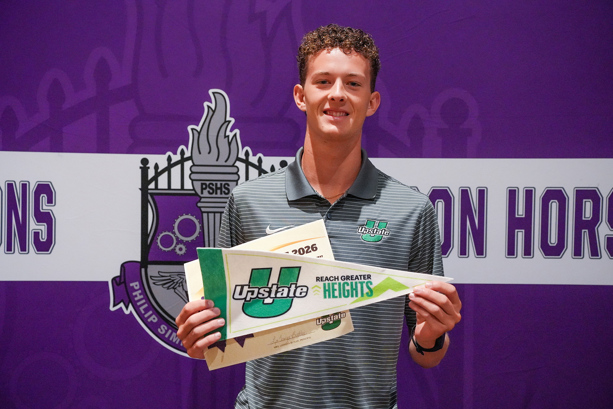 Man in gray polo holding pennant banner with school logo and text, standing before a purple backdrop with school crest.