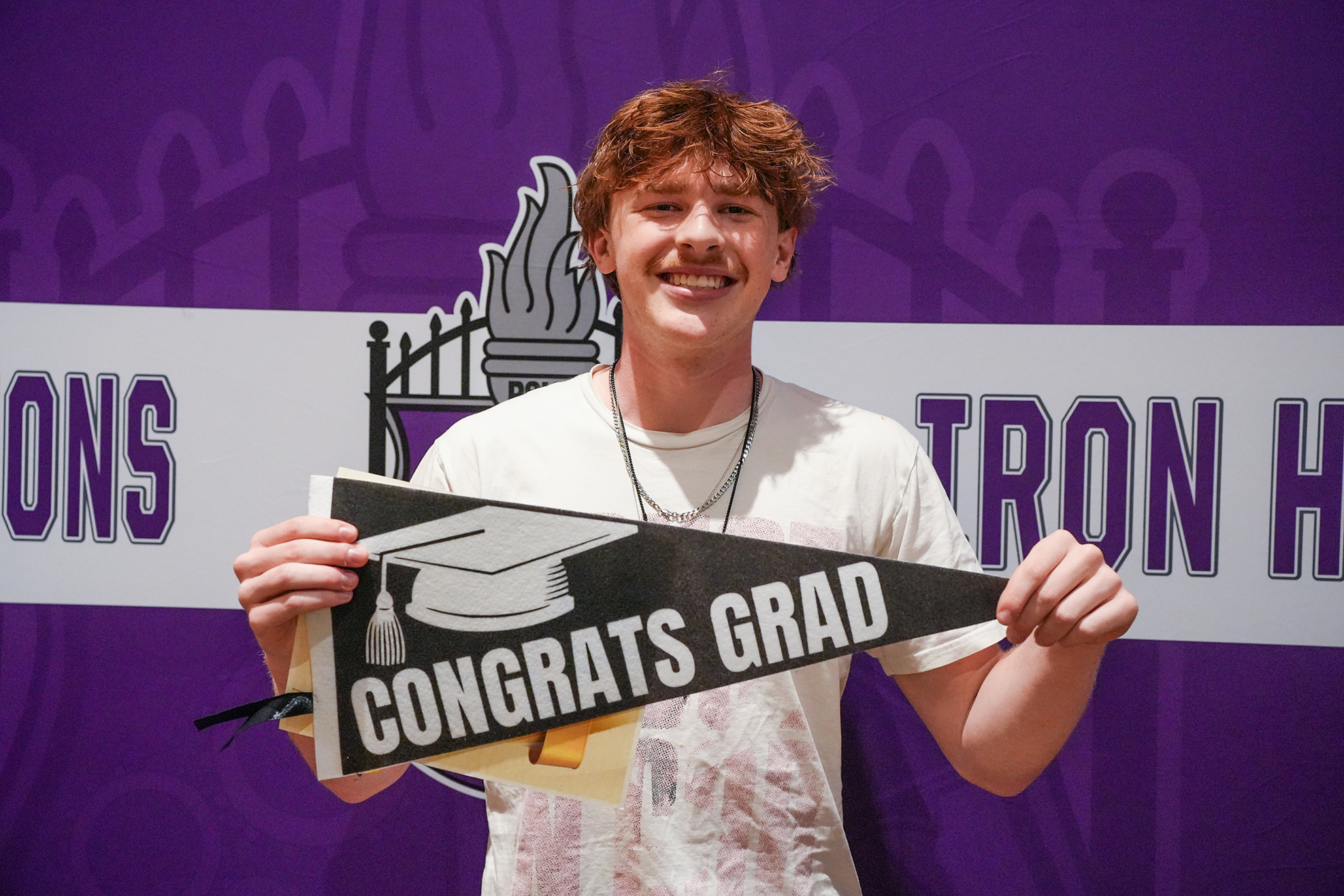 A smiling young man holds a graduation cap banner that says "CONGRATS GRAD" in front of a purple backdrop.