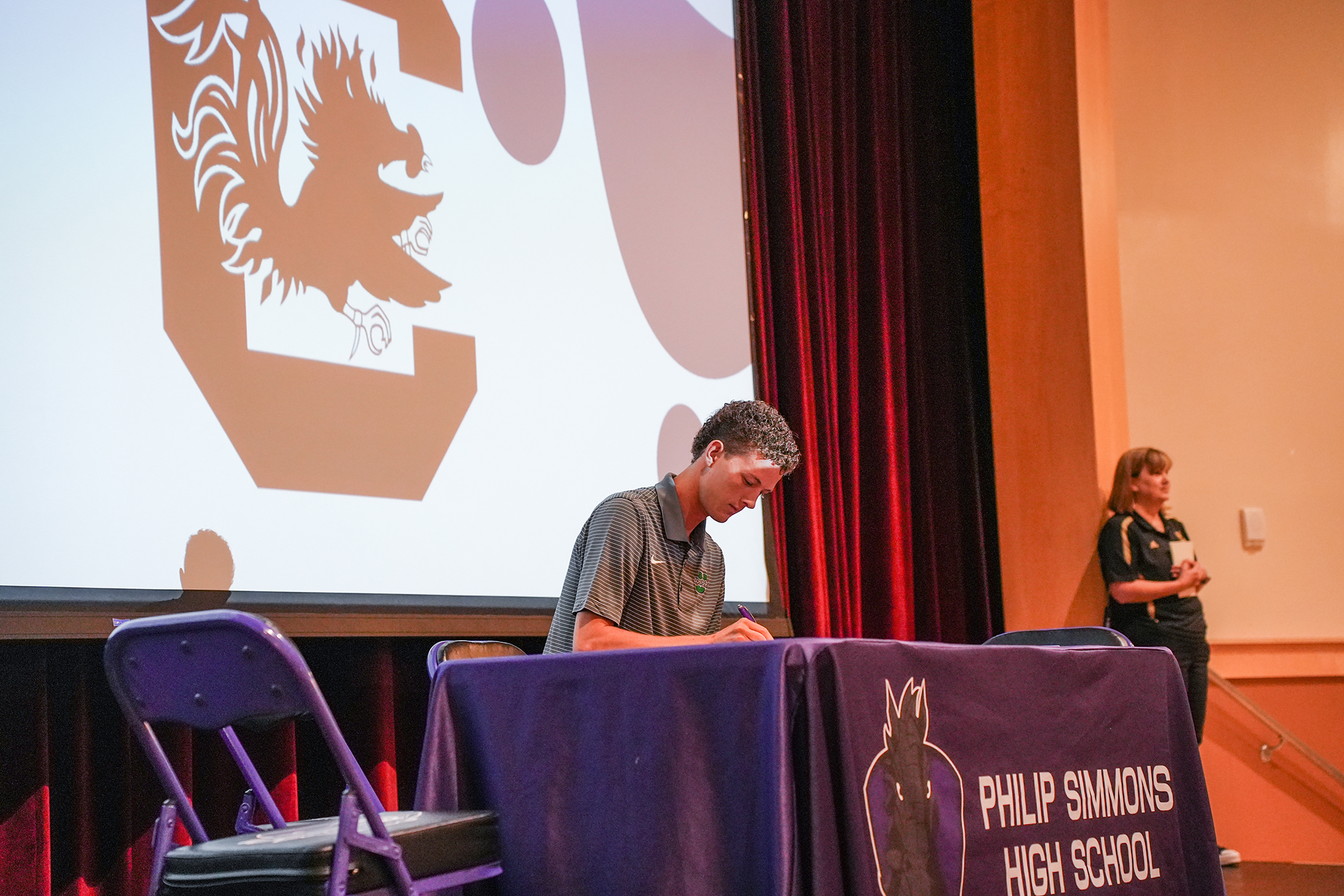 A man sits at a table with a purple tablecloth at a school event. Behind him, a projector screen shows the school logo.