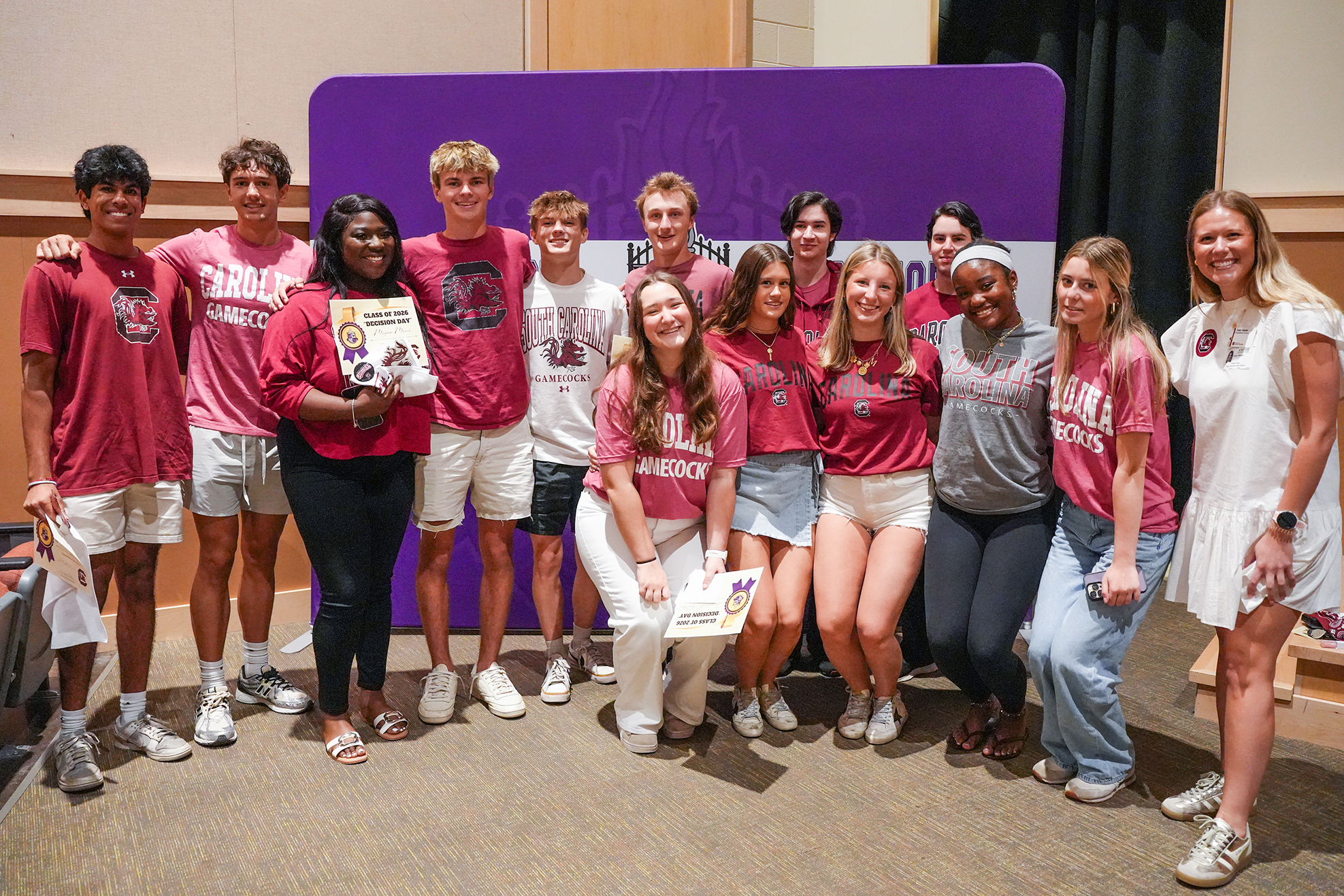 A group of students stands behind a purple backdrop, all wearing maroon shirts, except for one individual in gray.