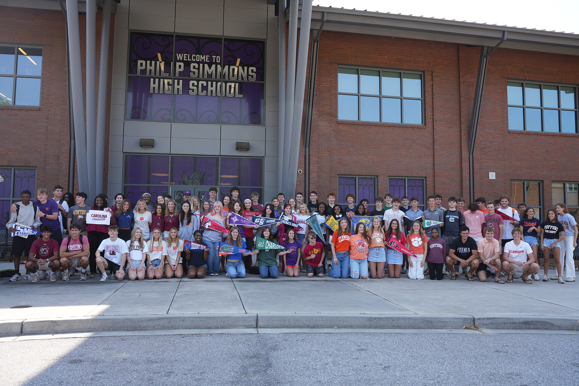A large group of students stands outside a brick school building with purple trim. A sign reads "Welcome to Philip Simmons High School."