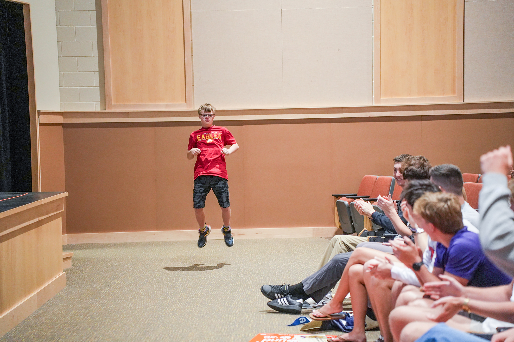 A boy jumps in mid-air wearing red t-shirt and black shorts, while others clap, seated on chairs in a room.