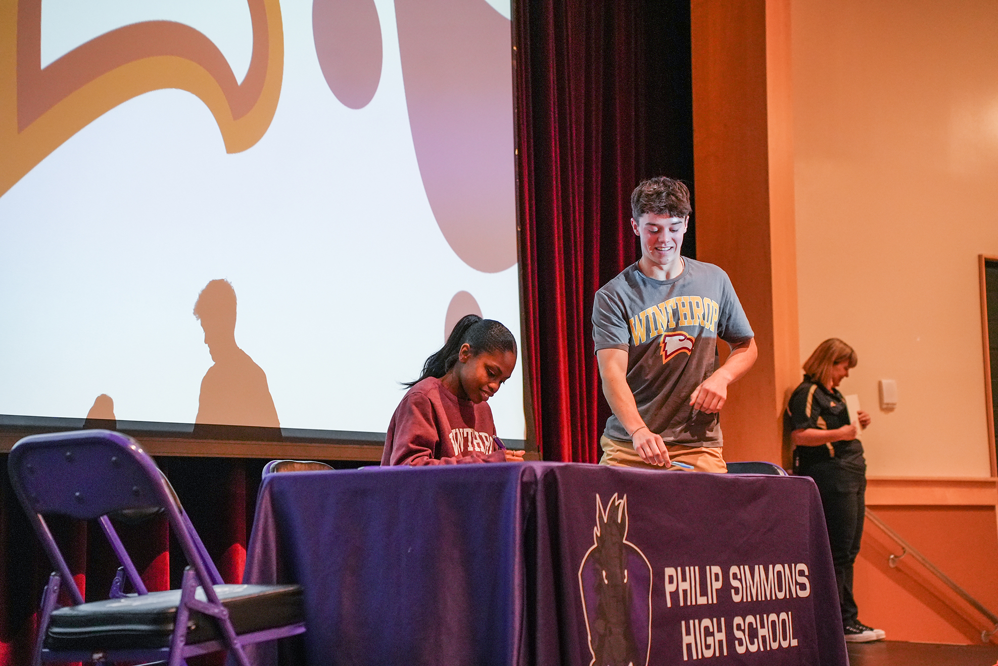 A stage with a backdrop shows two people at a table, a woman seated and a man standing.