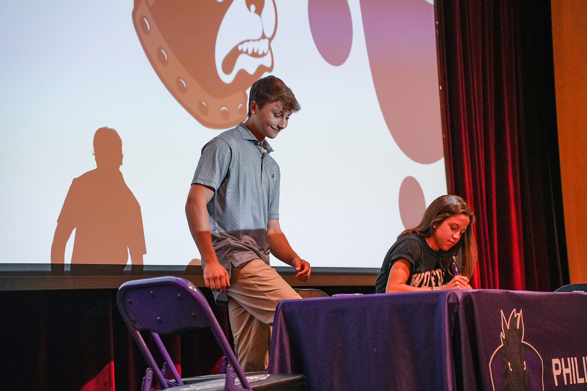 A man and a woman at a table with a purple tablecloth, projector screen showing a cartoon character.