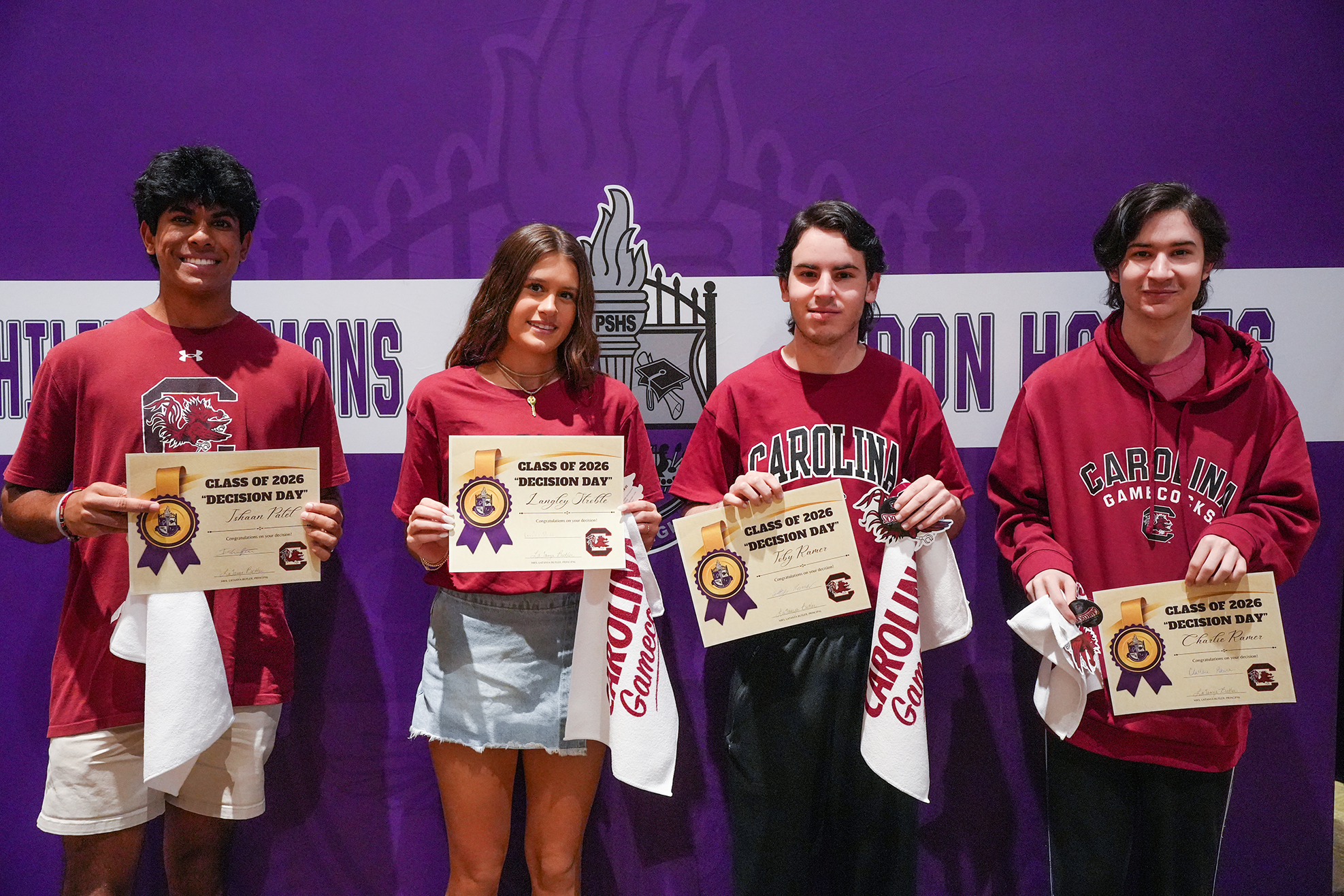 Four people in red shirts stand before a purple wall, holding certificates and towels. Two men wear white shorts and a red hoodie, while a woman wears a gray skirt.