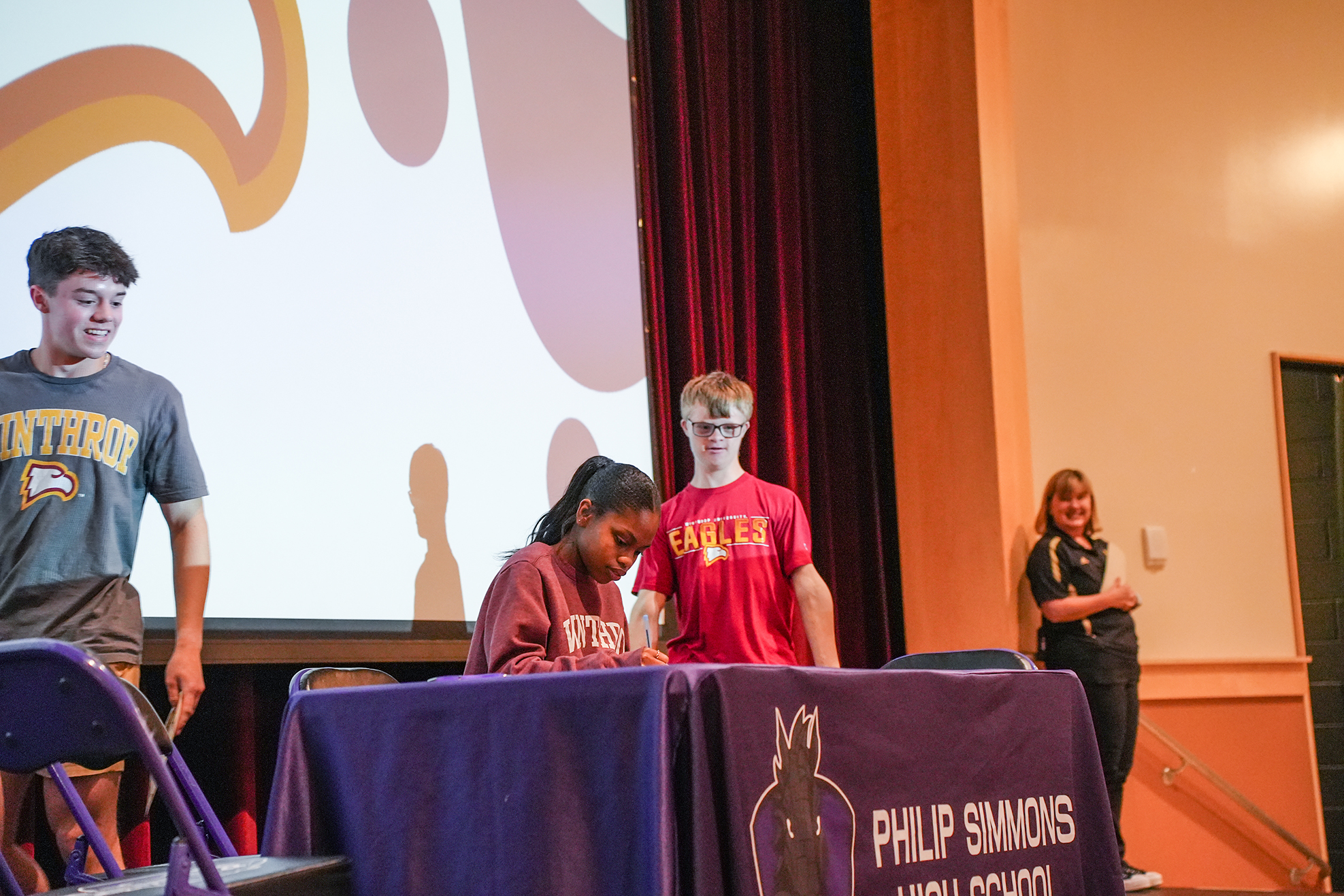 A presentation with a projector screen, featuring three individuals standing behind a table with a purple tablecloth.